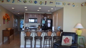 A kitchen with a central island, bar stools, and a dining area with a black leather chair and a lamp.