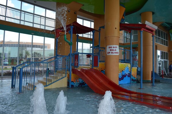 An indoor water park with a red slide, multiple water jets, and a 'Do Not Walk Up Slides' sign.