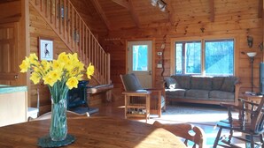 Interior de una cabaña de madera con una escalera, un sofá, una mesa con un jarrón de flores y un televisor.