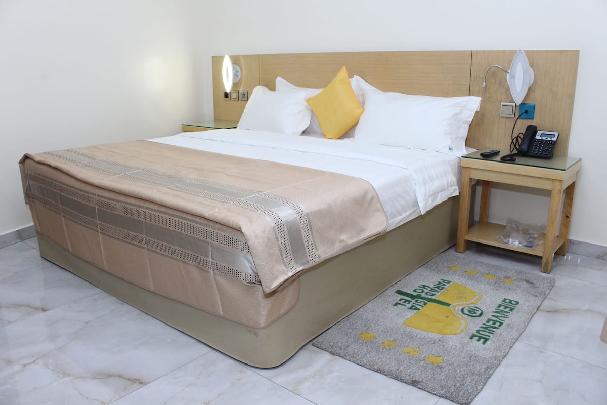 A neatly made bed with a beige bedspread and pillows, a bedside table with a lamp and phone, and a welcome mat with a logo.