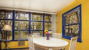 Une pièce avec une table ronde blanche, quatre chaises blanches, un vase violet avec des fleurs et de grandes fenêtres aux montants bleus.