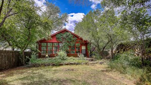 Une maison en bois rouge, dotée de grandes fenêtres et d’un porche, entourée d’arbres et d’une cour verdoyante.