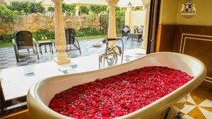 Una bañera llena de pétalos de rosa roja, un suelo a cuadros y vistas de un jardín con sillas y una fuente.