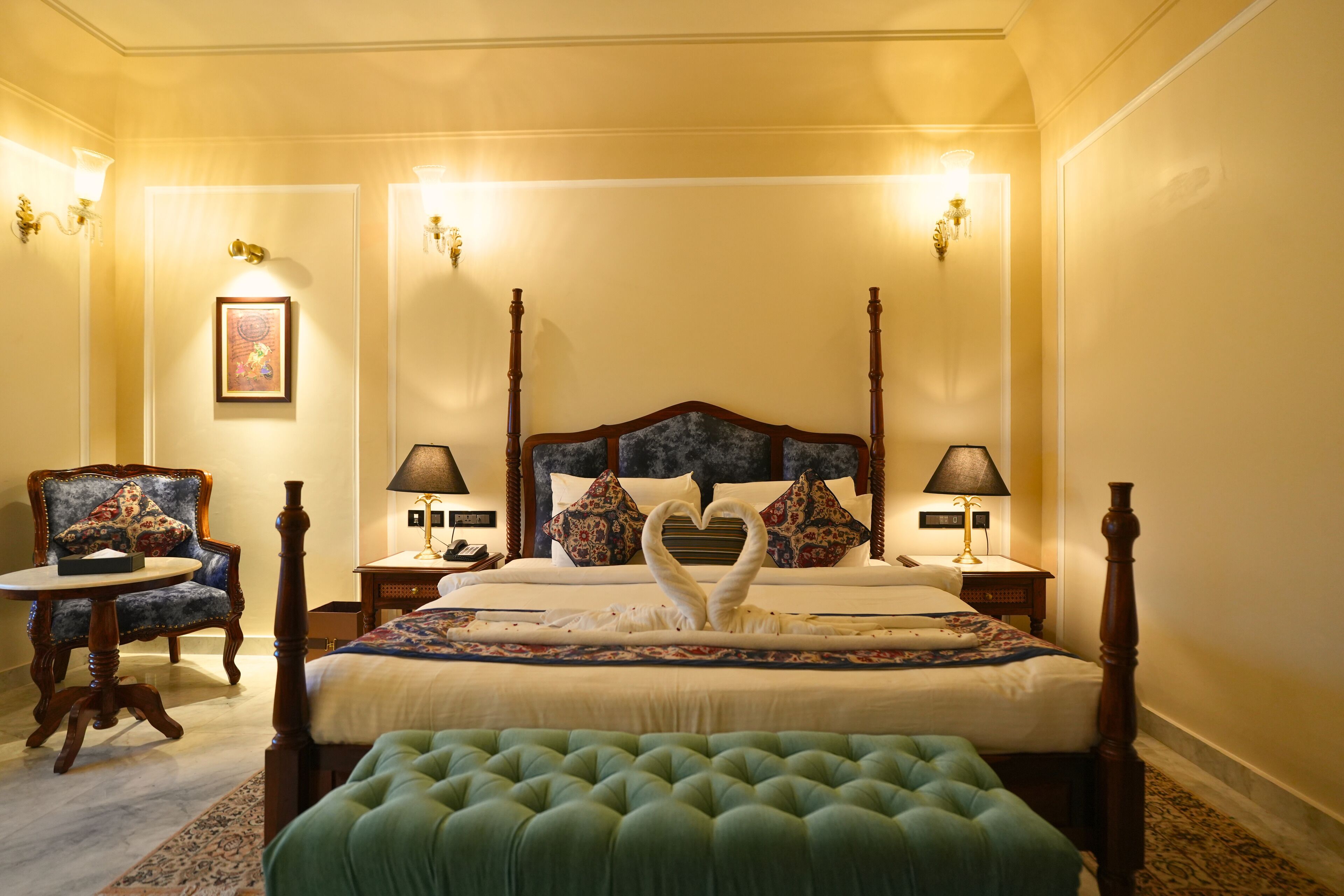 Una cama con dosel adornada con una decoración de cisne en forma de corazón, un banco tapizado en verde y un sillón clásico.