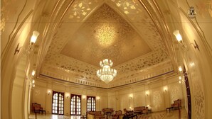 Un salón imponente con techo ornamentado y candelabros, disposición de asientos y ventanas amplias.