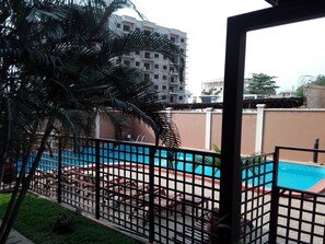 A view of a swimming pool area with a fenced deck, palm trees, and a building in the background. - Trevi Towers (Accra)