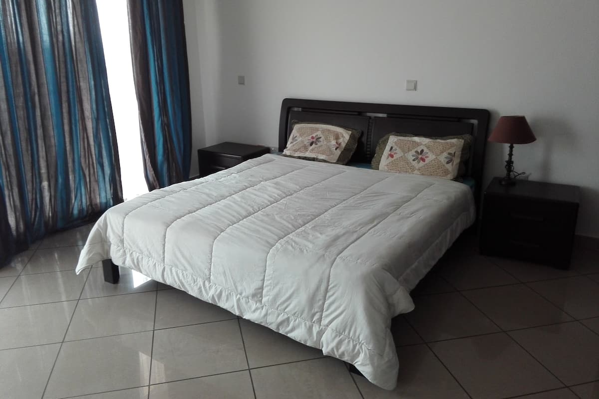 Una cama bien tendida con edredón blanco, dos mesitas de noche con lámparas y una ventana con cortinas azules.