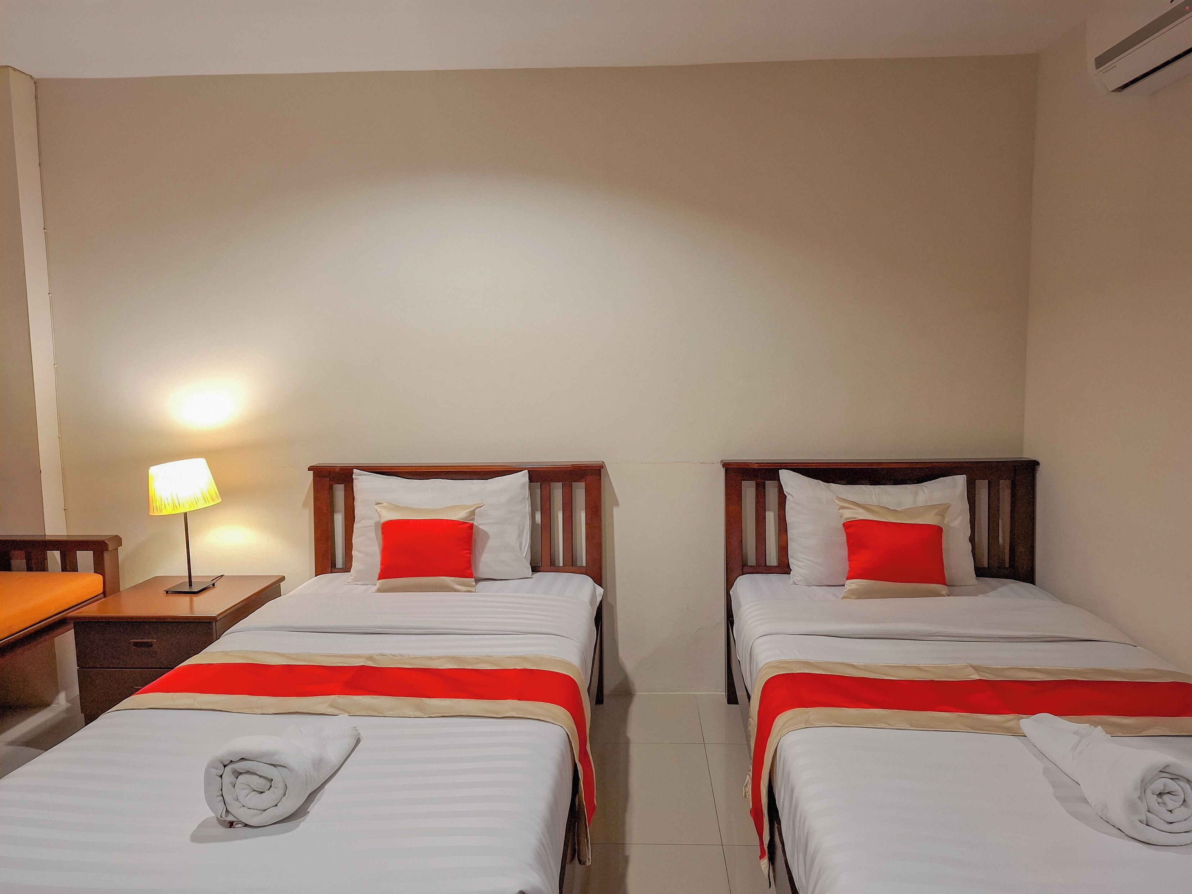 A hotel room with two single beds, each with a red and white pillow, a wooden headboard, and a red and white bedspread. There is a nightstand with a lamp and a folded towel on the bed.