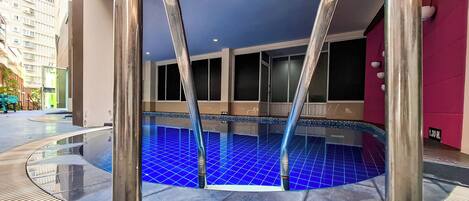 A modern indoor swimming pool with clear blue tiles, surrounded by a wooden deck and metal railings.