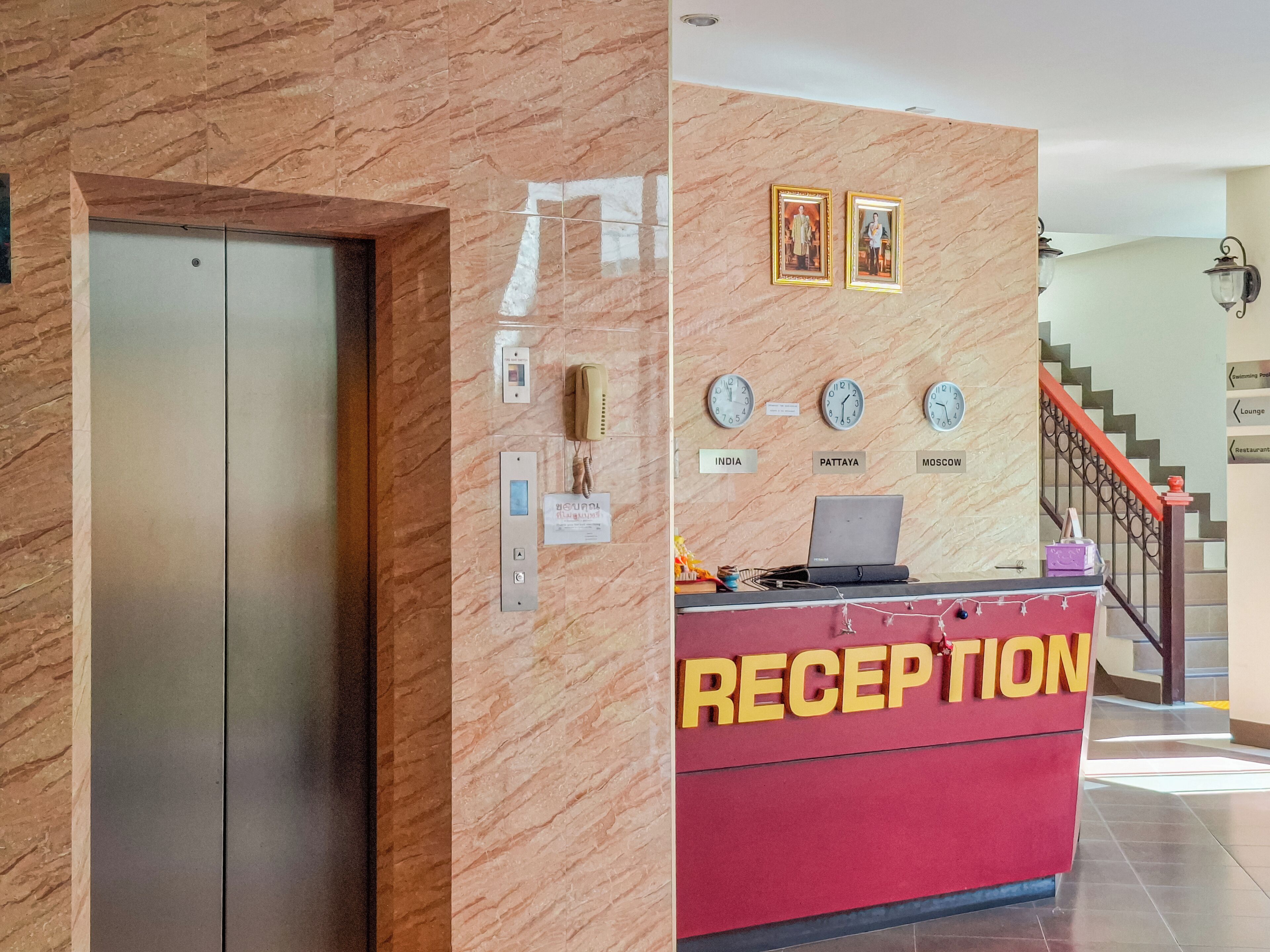 Mostrador de recepción con un cartel que dice 'RECEPTION', un teléfono y relojes que marcan 'INDIA', 'PATTAYA' y 'MOSCOW'.