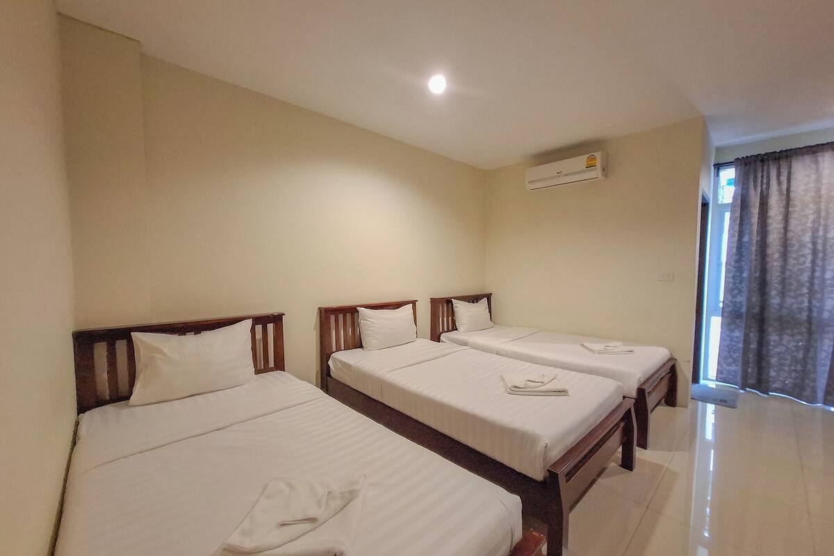 A hotel room with three single beds, a window with curtains, and an air conditioning unit on the wall.