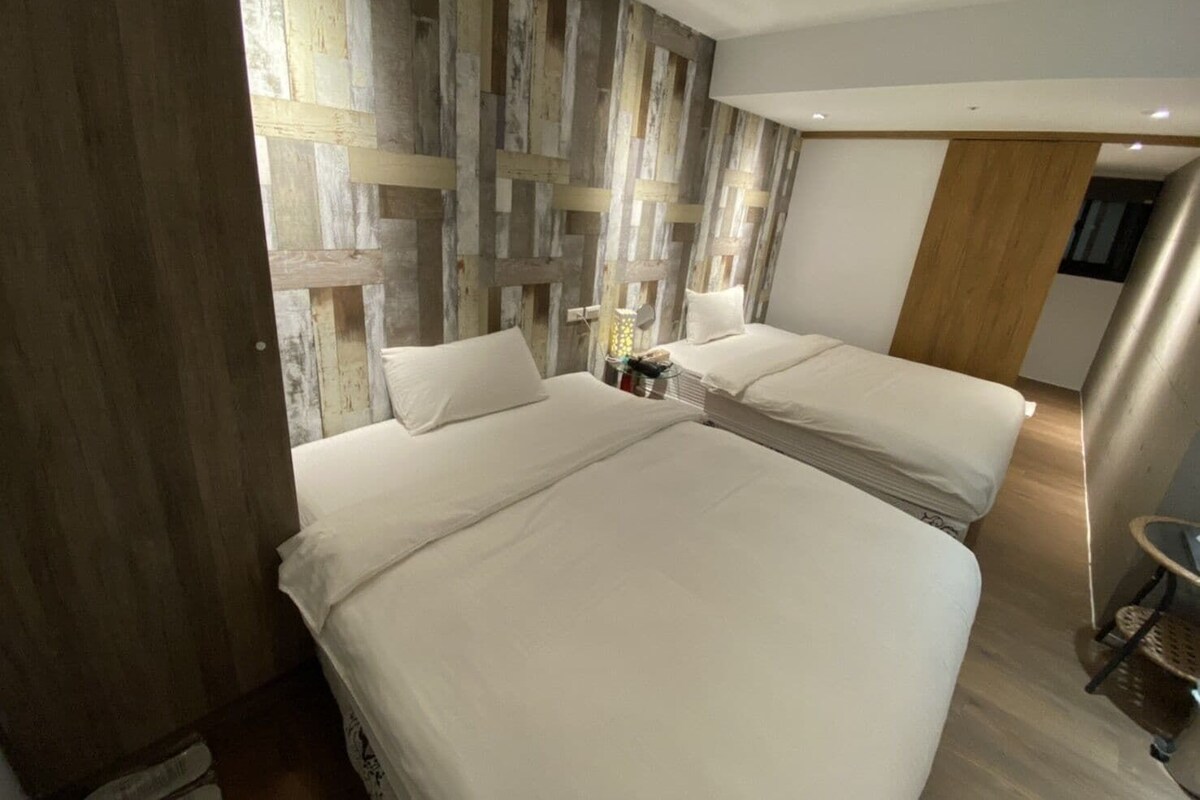 A hotel room with two beds, a wooden accent wall, a small table, and a door.