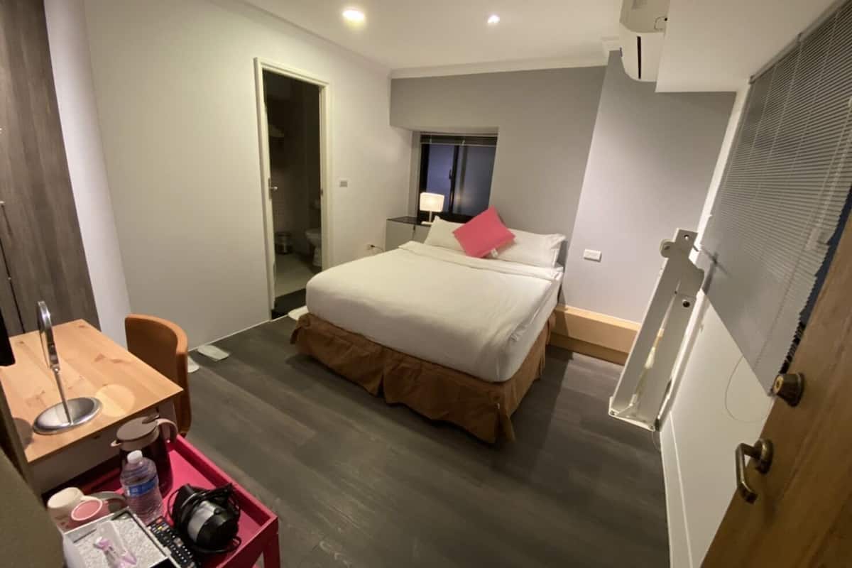 A hotel room with a bed, a desk with a chair, a small table, and a trash bin.