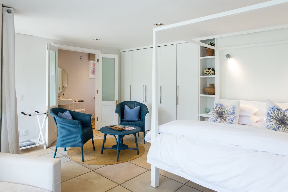 A bedroom with a bed, two chairs, a small table, a wardrobe, and a bathroom visible in the background.