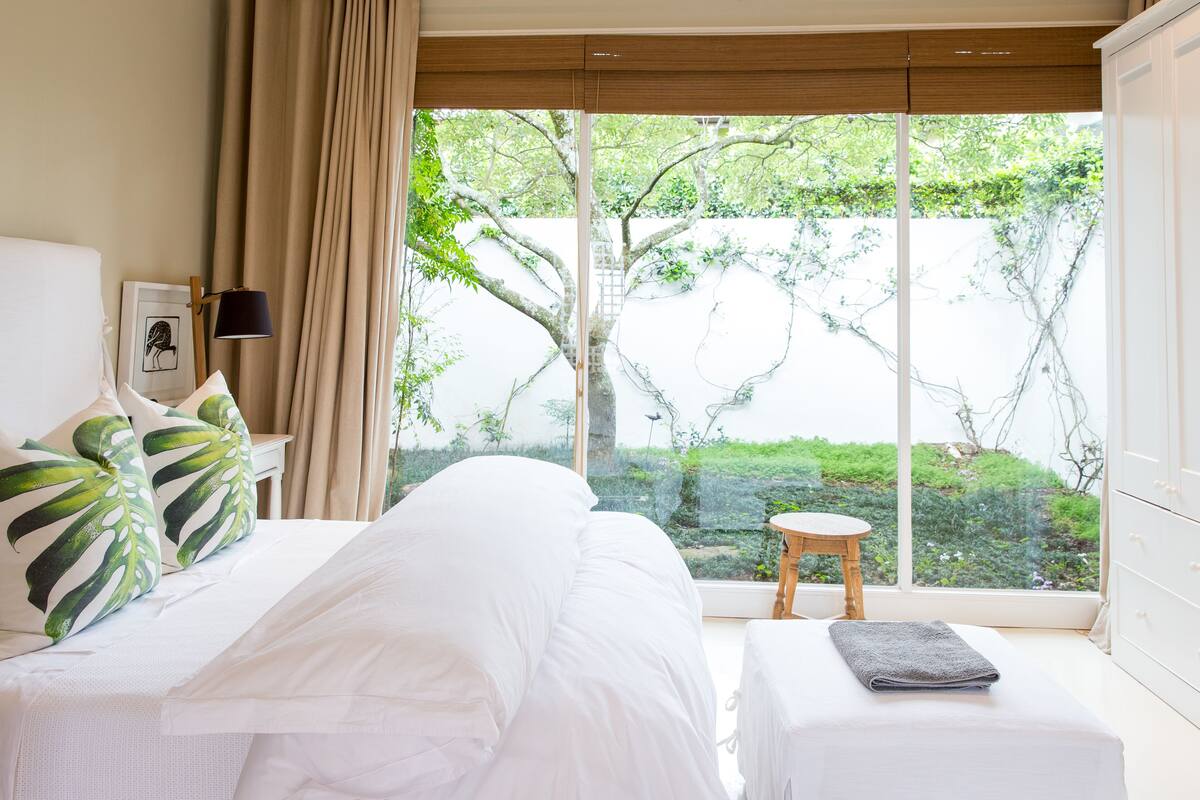 A bedroom with a large window, a bed, a bedside table, and a view of greenery outside.