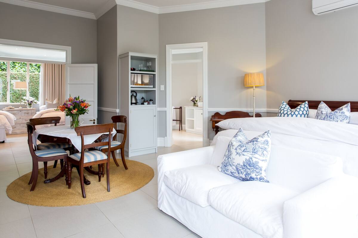 A hotel room with a large bed, a dining area with a table and chairs, a wardrobe, and a view of the outdoors.