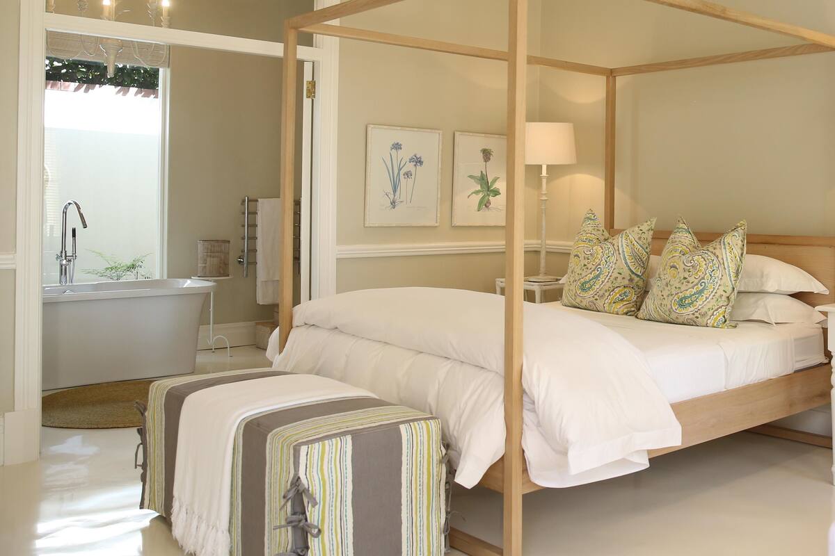 A four-poster bed with white bedding, a bedside table with a lamp, and two framed botanical prints on the wall.