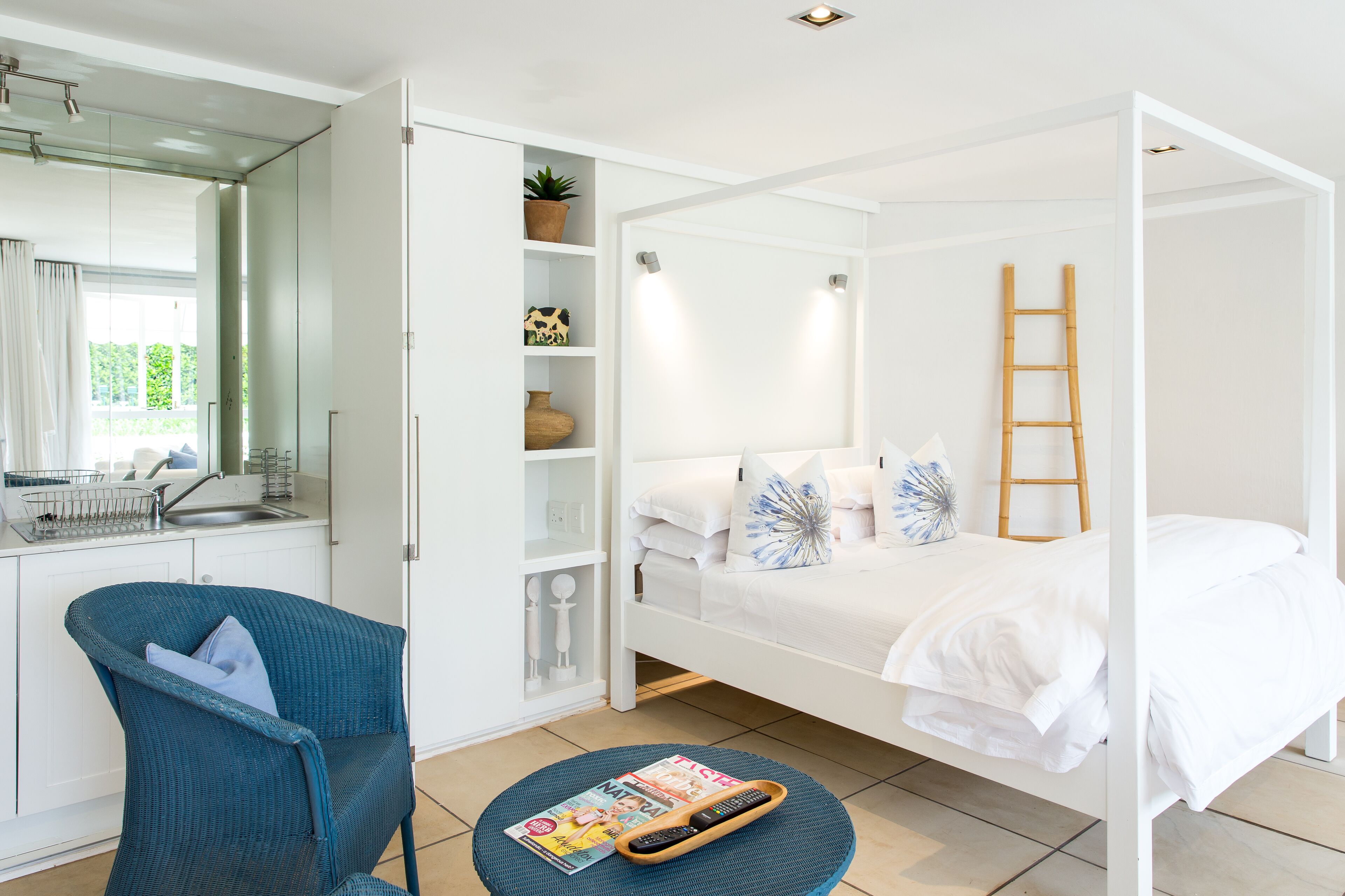 A modern bedroom with a white bed, a blue chair, a round table with a magazine, and a kitchen area with a sink and cabinets.
