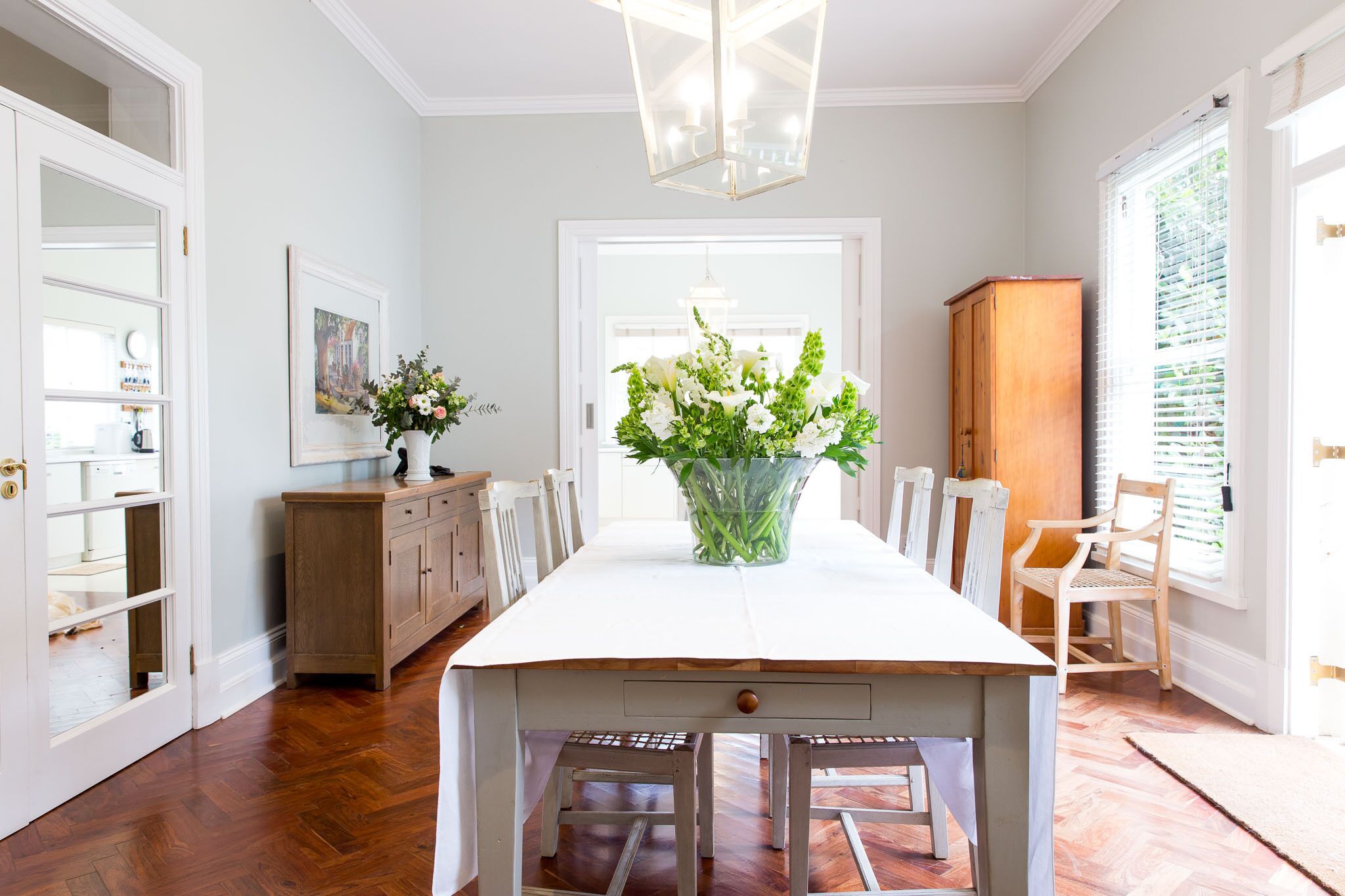 A dining room with a long white table, wooden chairs, a cabinet, and a vase of flowers.