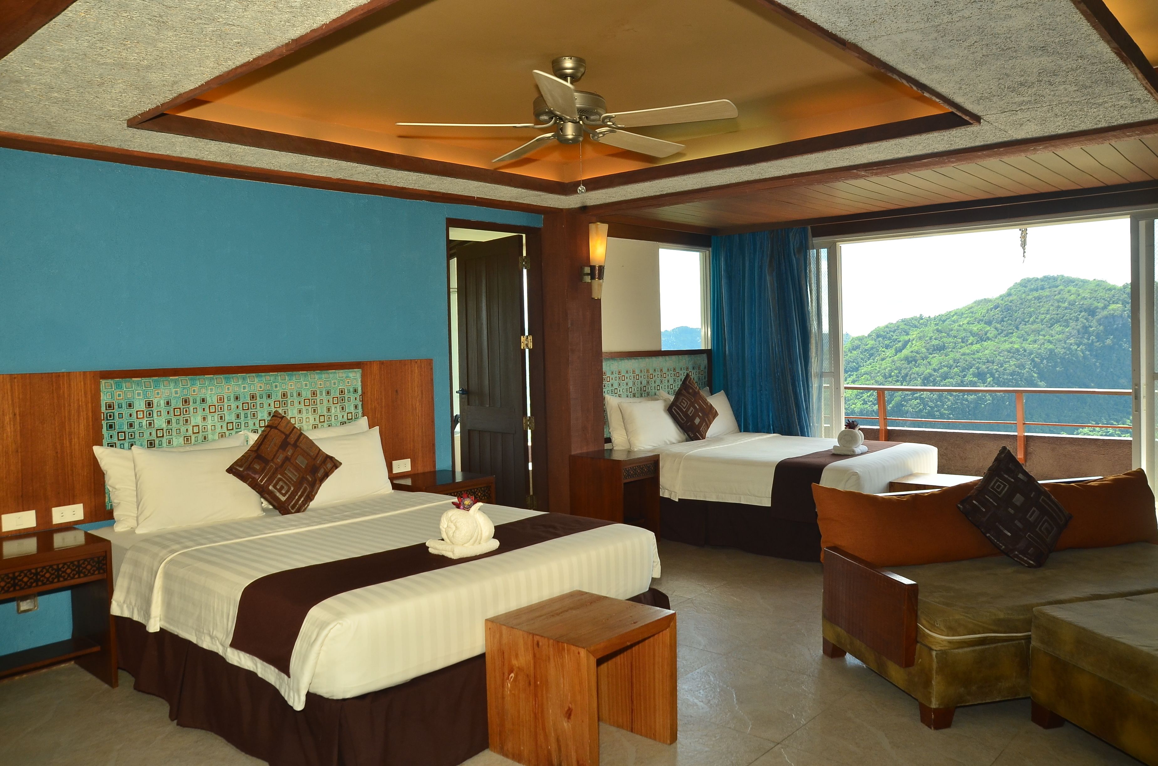 A hotel room with two beds, a ceiling fan, a large window with a view of a mountain, and a wooden headboard.