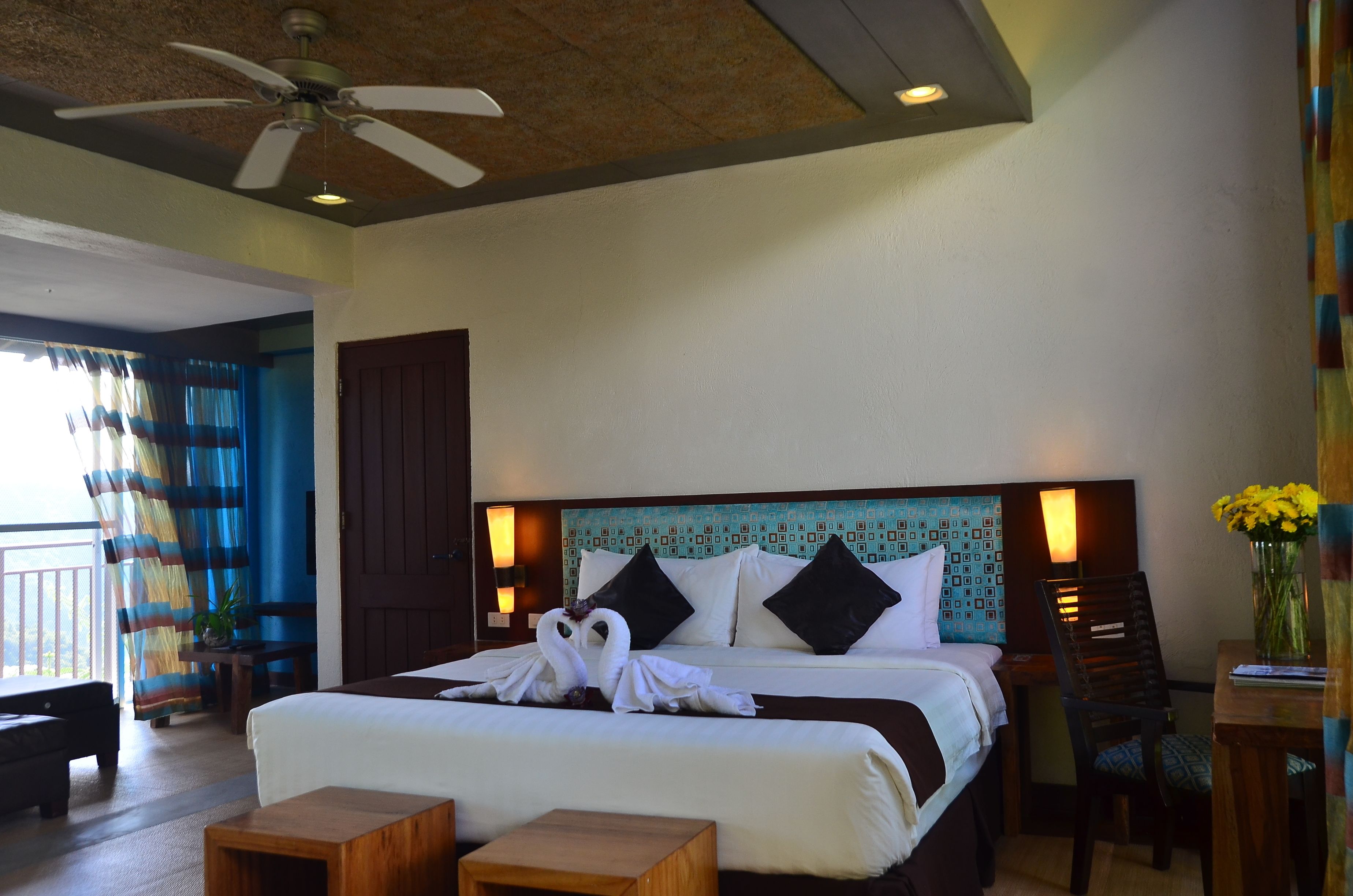A hotel room with a bed, a desk, a chair, a ceiling fan, and a view of the outdoors.