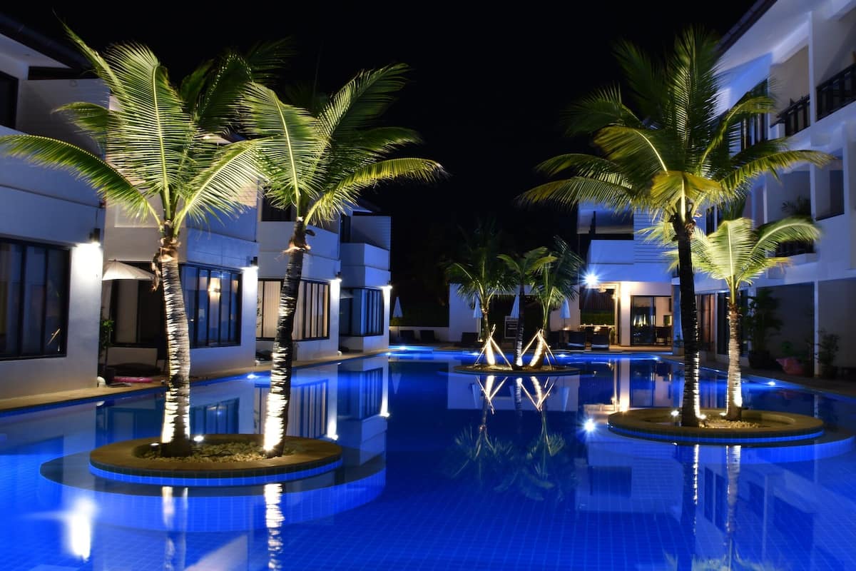 A swimming pool with palm trees and illuminated edges, surrounded by modern buildings with balconies.