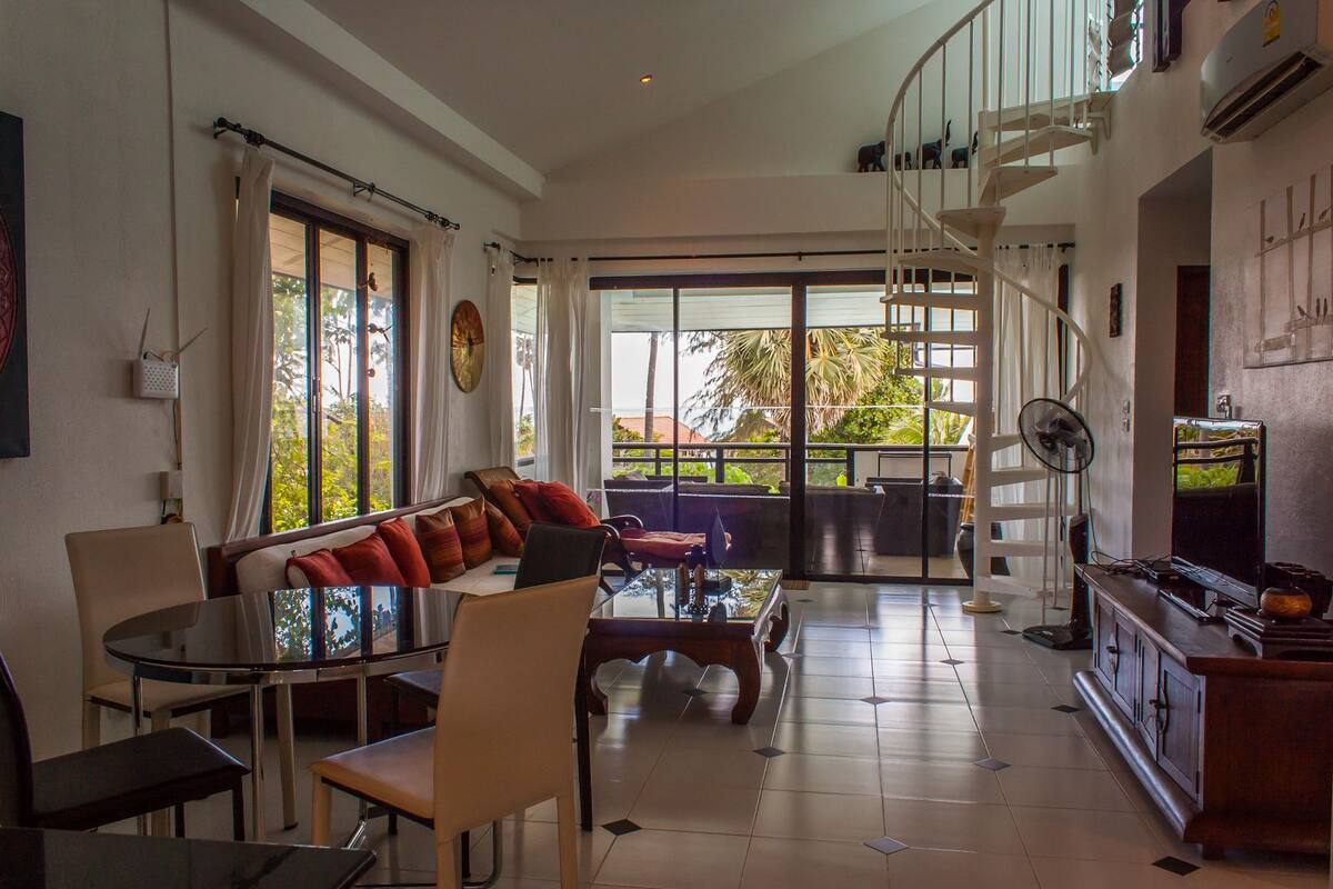 A spacious living area with a glass coffee table, a sofa with red cushions, a dining table with chairs, and a staircase leading to an upper level.