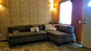 Un salón con un sofá modular gris oscuro, cojines estampados, suelo de madera y una pared con estampado geométrico.