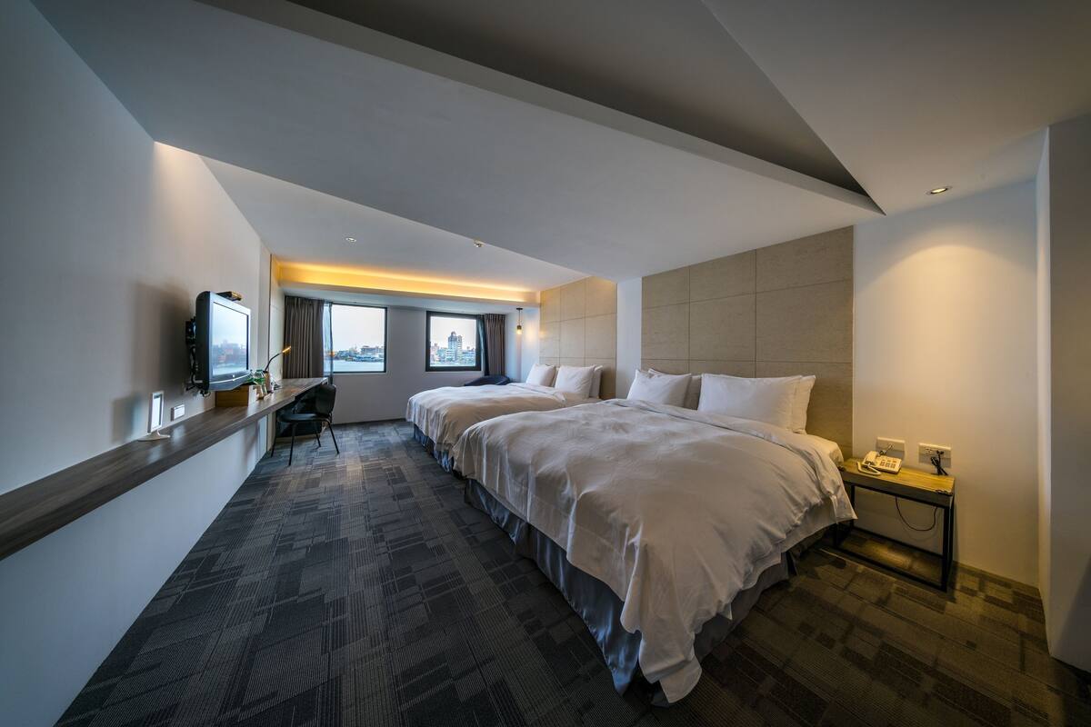Ein Hotelzimmer mit einem großen Bett, einem Schreibtisch mit Stuhl, einem Fernseher und Blick auf die Stadt durch das Fenster.