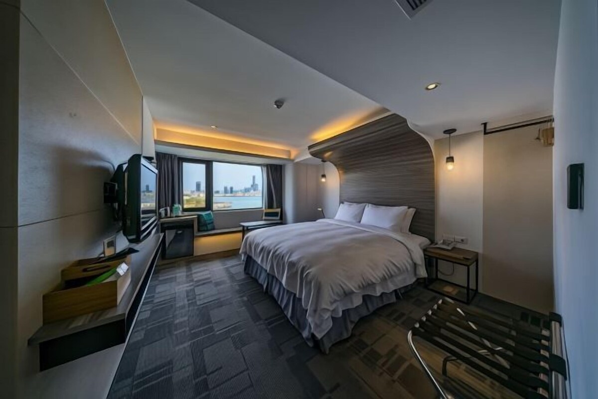 A modern hotel room with a large bed, a TV, a desk, and a city view.