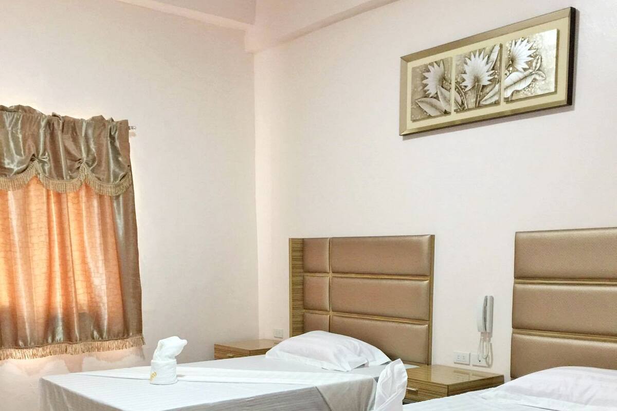 Hotellihuone, jossa on kaksi erillistä sänkyä, puiset päätylevyt, yöpöytä lampun kanssa ja seinällä oleva kehystetty, kukkateemainen taideteos.
