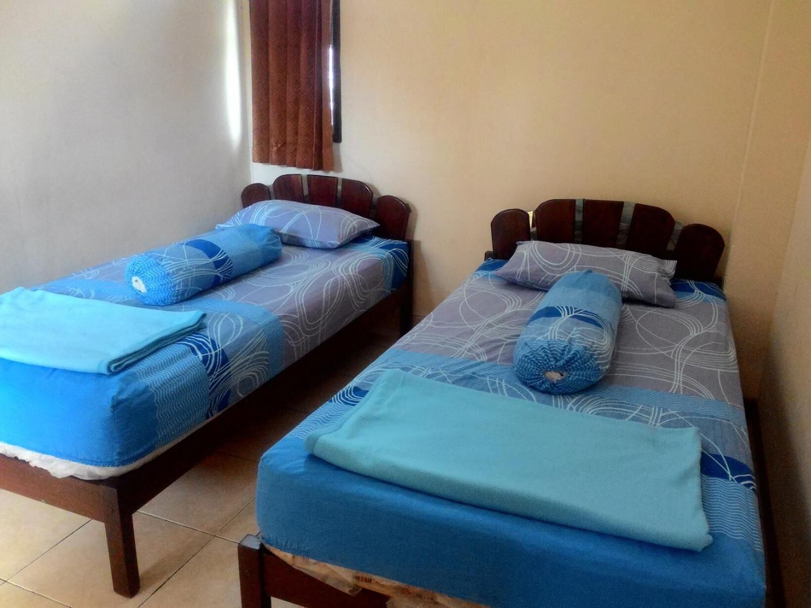 Dos camas individuales con ropa de cama azul y cabeceros de madera en una habitación con suelo de baldosas.