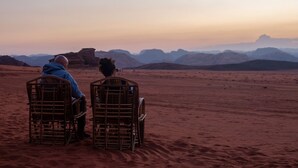 Two people sitting on wicker chairs facing a desert landscape with mountains in the distance. - Desert Moon Camp (Wadi Rum)