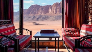 A room with a large window offering a view of a desert landscape. There are two sofas with patterned cushions and a small table in the center. - Desert Moon Camp (Wadi Rum)