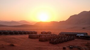 A desert landscape with a sunset, featuring several wooden cabins with striped awnings. - Desert Moon Camp (Wadi Rum)