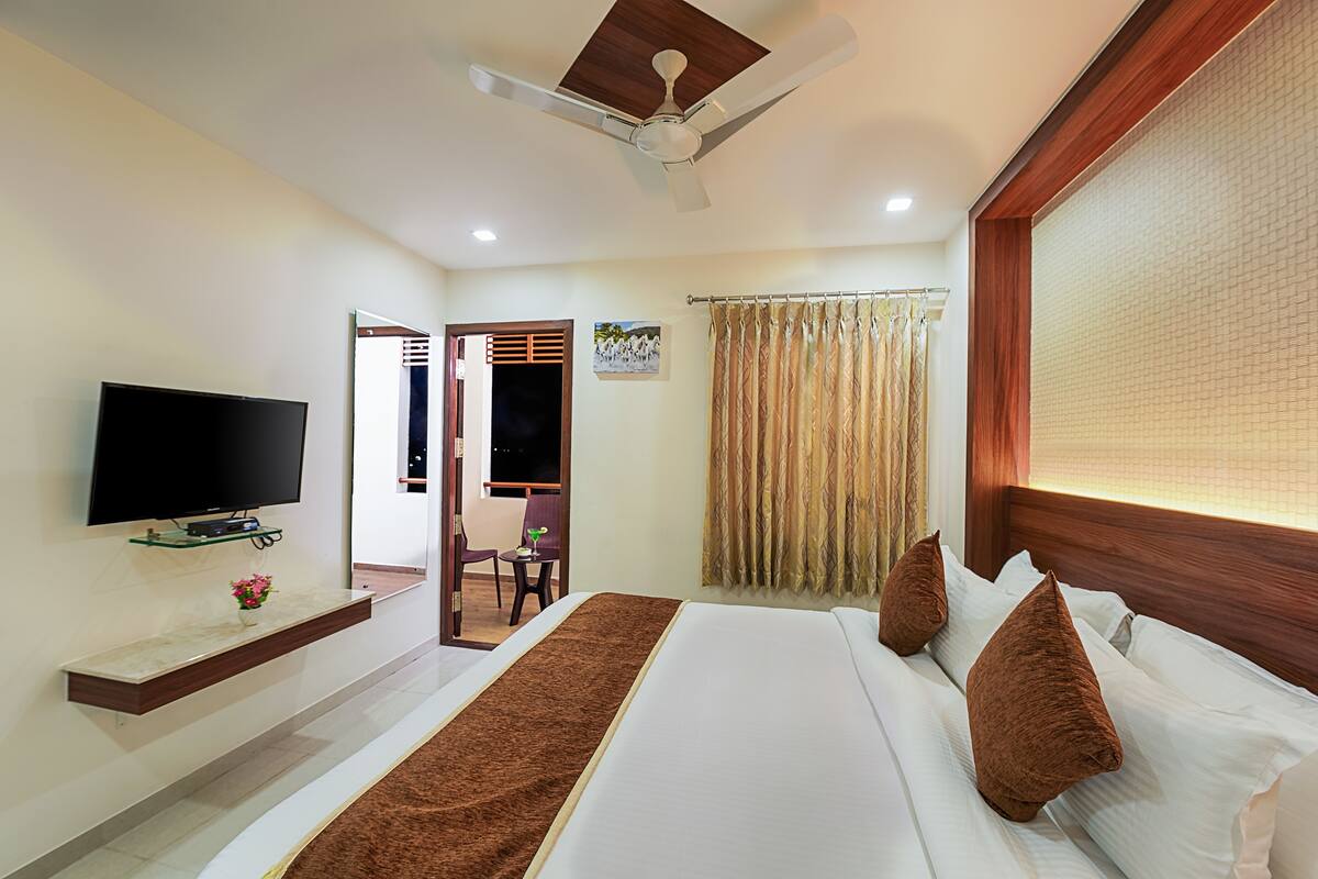 A hotel room with a bed, a TV mounted on the wall, a ceiling fan, and a window with curtains.