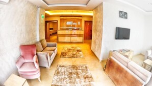Un salon moderne avec un canapé, des fauteuils et un téléviseur. On aperçoit à l’arrière-plan un coin cuisine avec des placards et un plan de travail.
