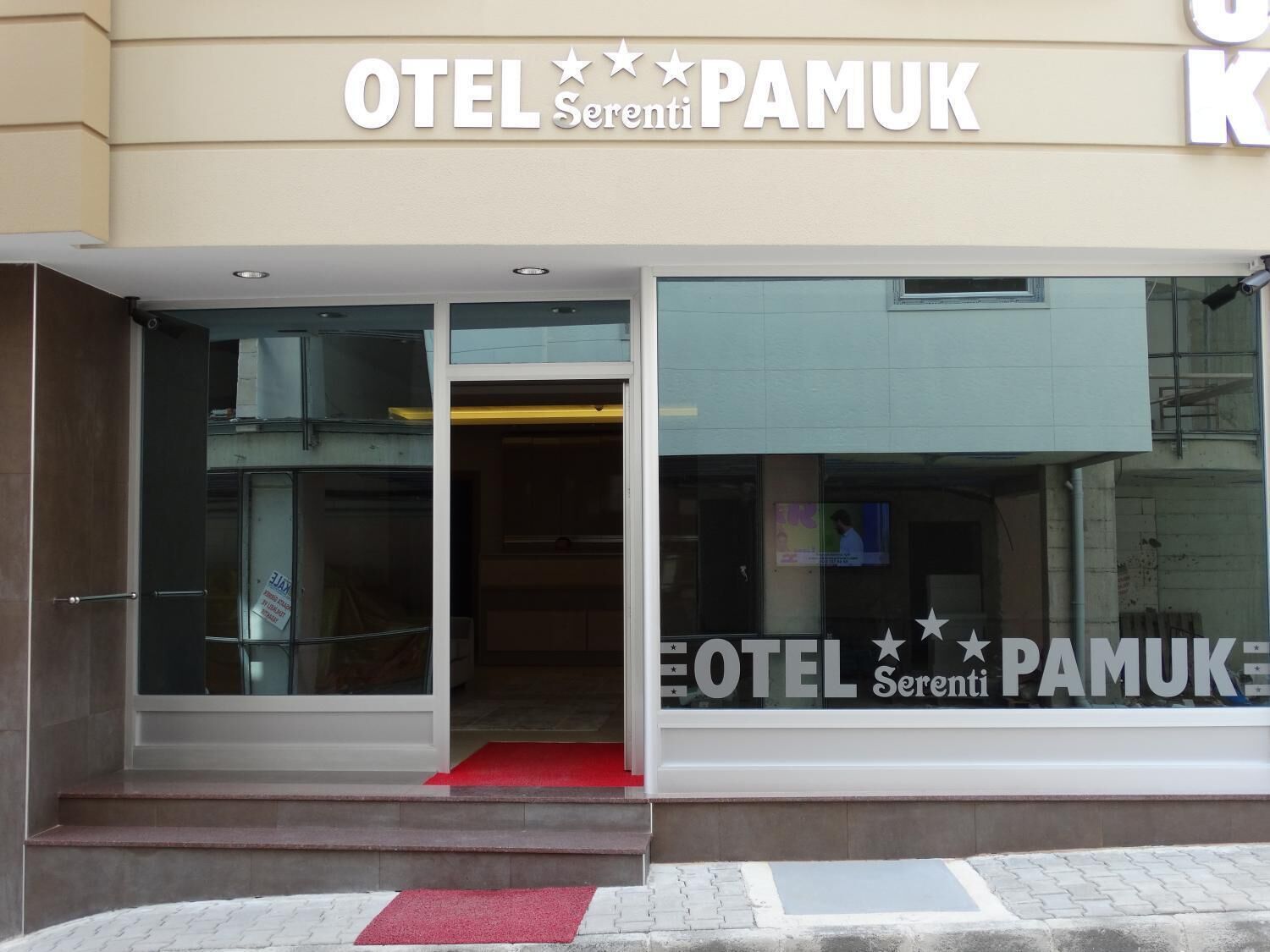 Ingången till ett hotell som heter ”OTEL Serenti PAMUK”, med en glasfasad, en röd matta och en synlig trappa som leder upp till entrén.