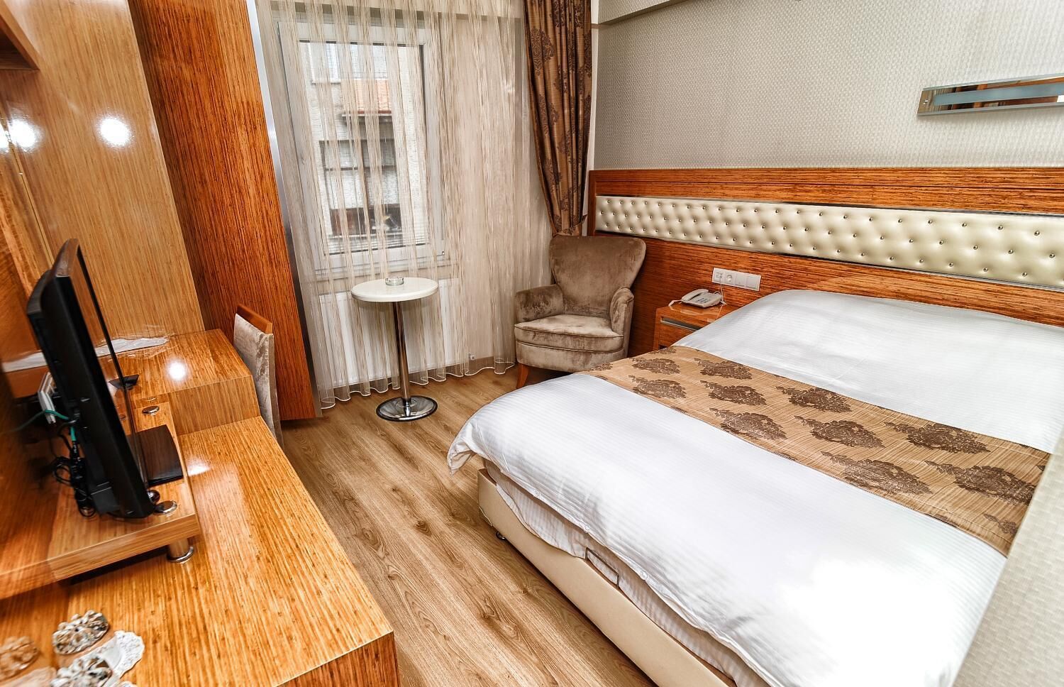 Una habitación de hotel con piso de madera, una cama con cabecera capitonada, una mesita redonda pequeña, una silla y un televisor sobre un mueble de madera.