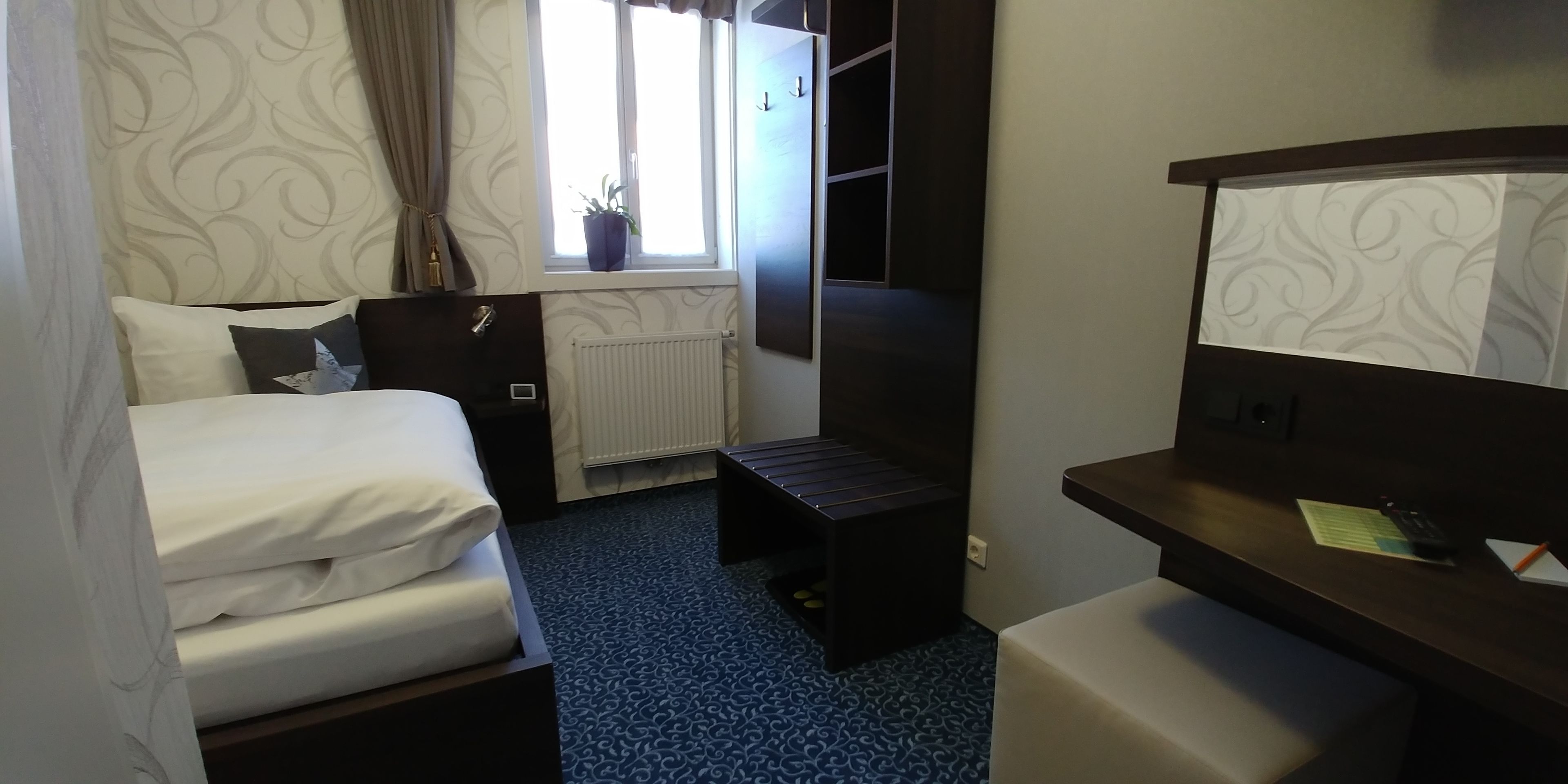 A hotel room with a bed, a desk, a chair, a radiator, and a window with curtains.