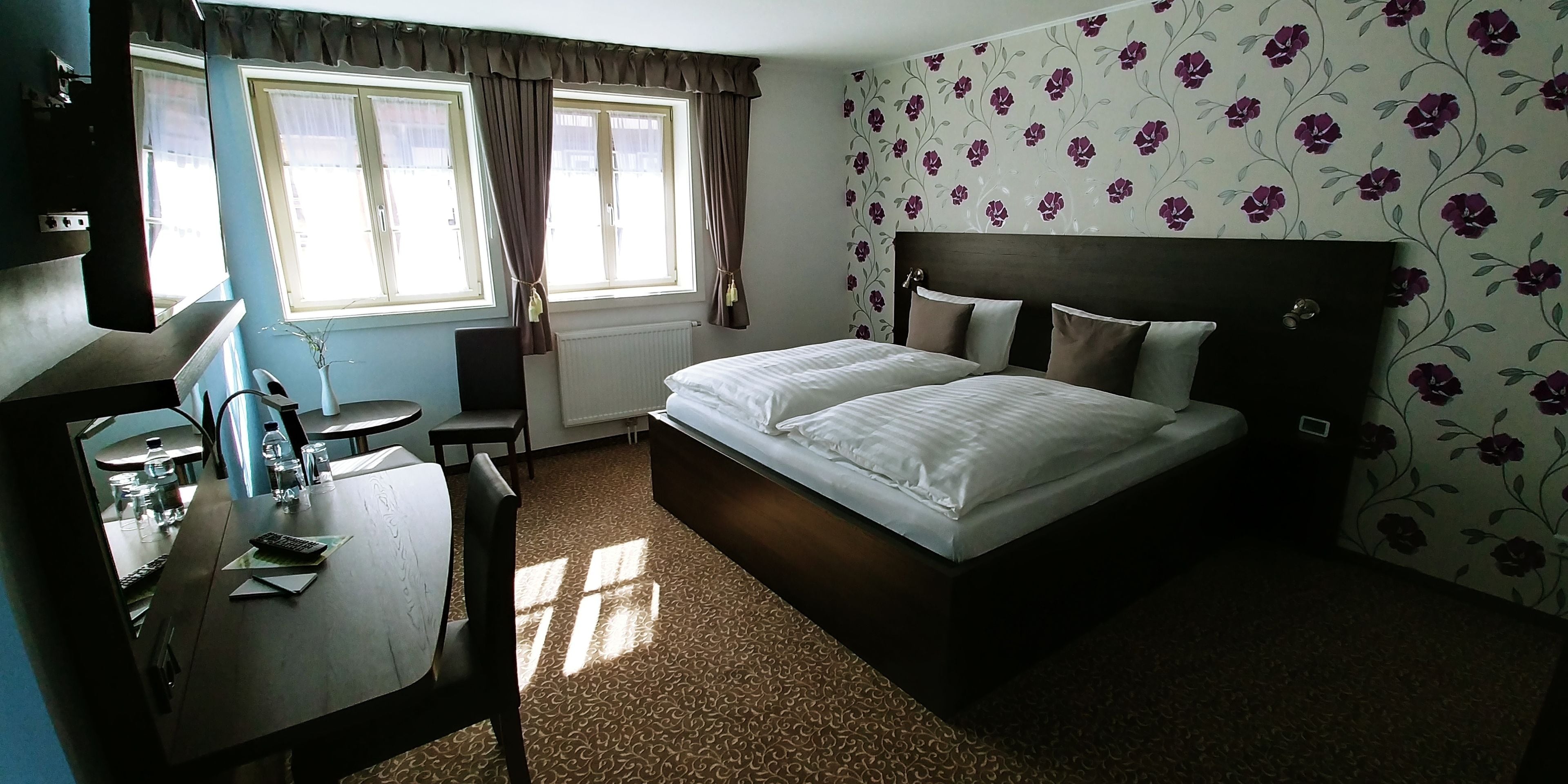 A hotel room with a bed, a desk with a chair, a window with curtains, and a floral wallpaper.