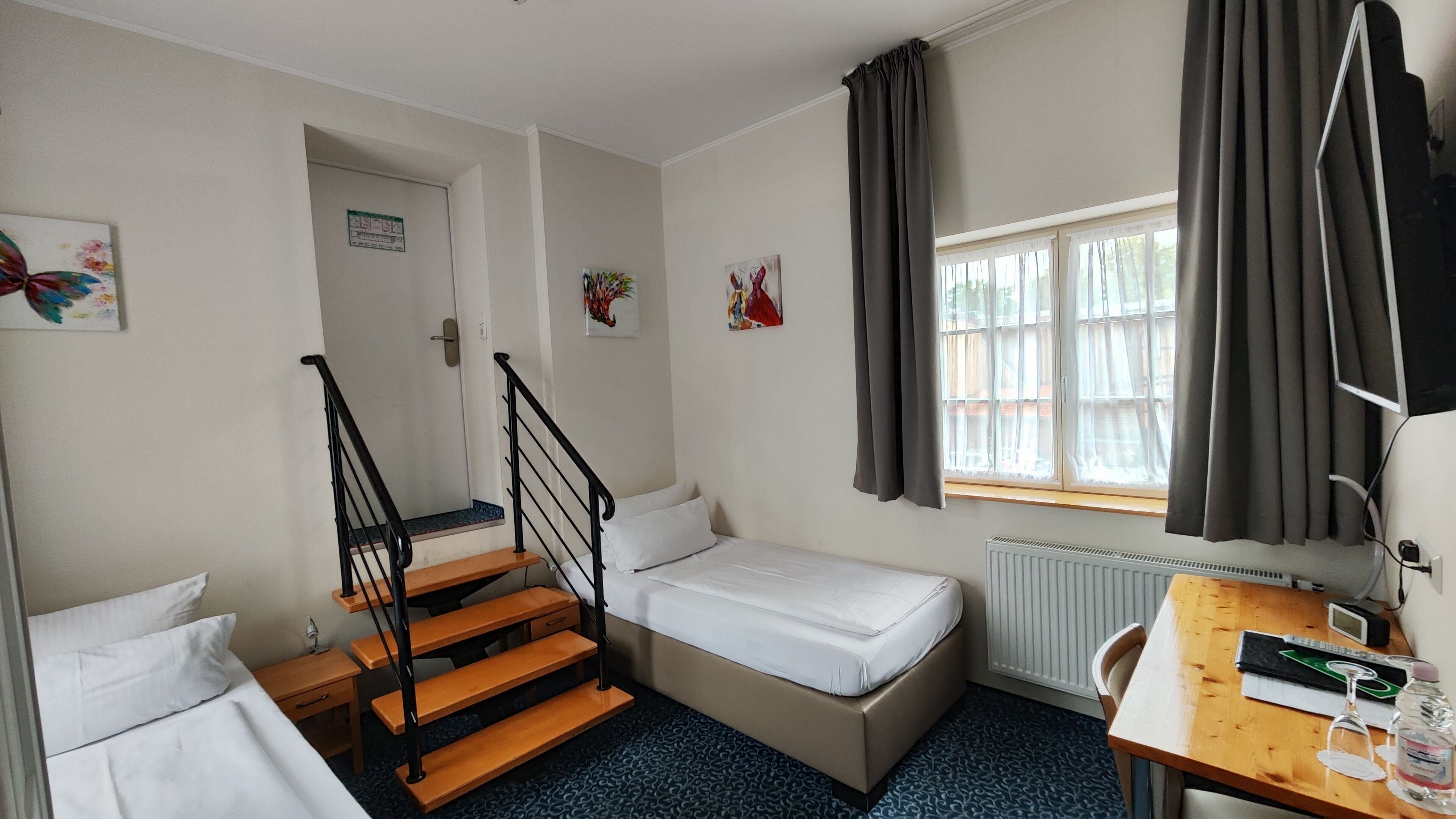 Habitación compacta con cama, escritorio con ordenador, ventana con cortinas, radiador y una escalera.