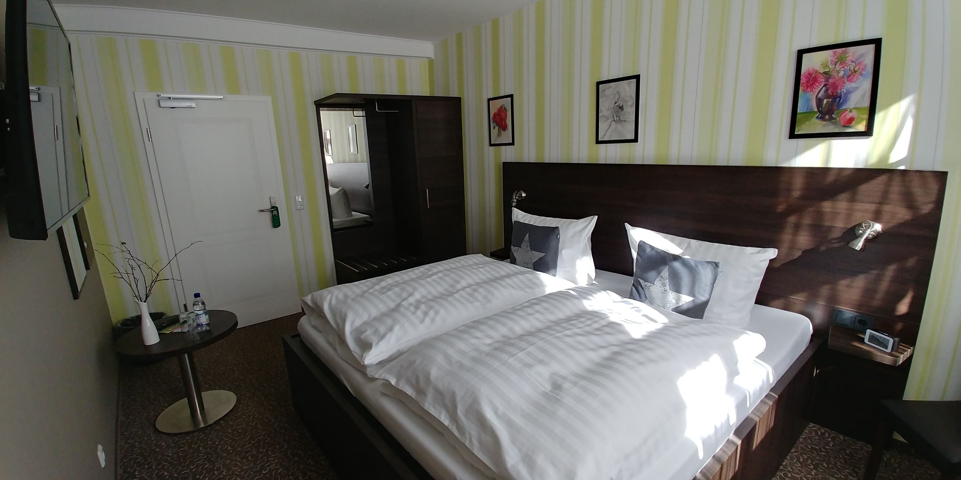 Hotelkamer met een groot bed, nachtkastje, spiegel, kledingkast en een klein rond tafeltje met een vaas en fles.