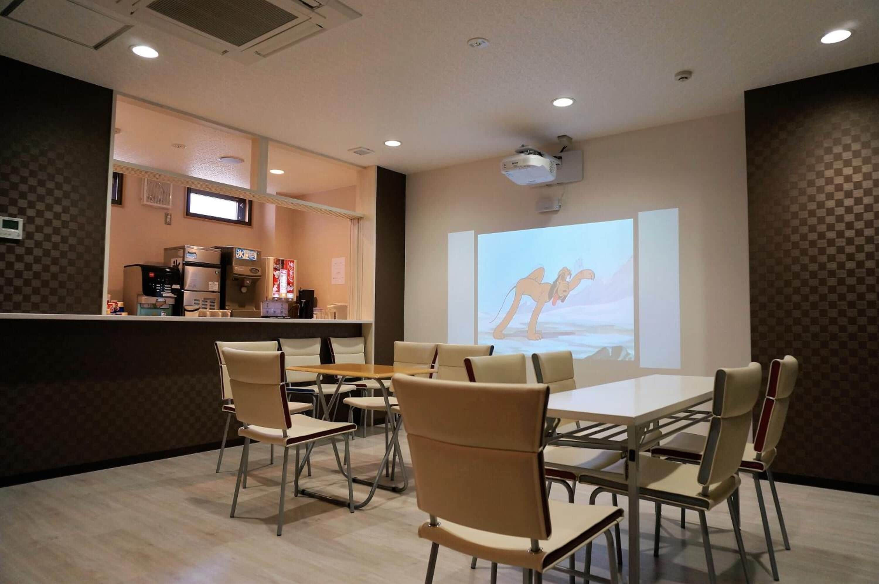 Een vergaderzaal met een grote tafel, stoelen, een projectorscherm en een kitchenette op de achtergrond.