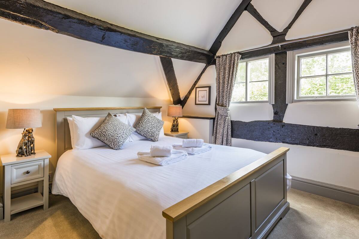 A neatly made bed with pillows and towels, a bedside lamp, and a nightstand with a lamp and a decorative item.