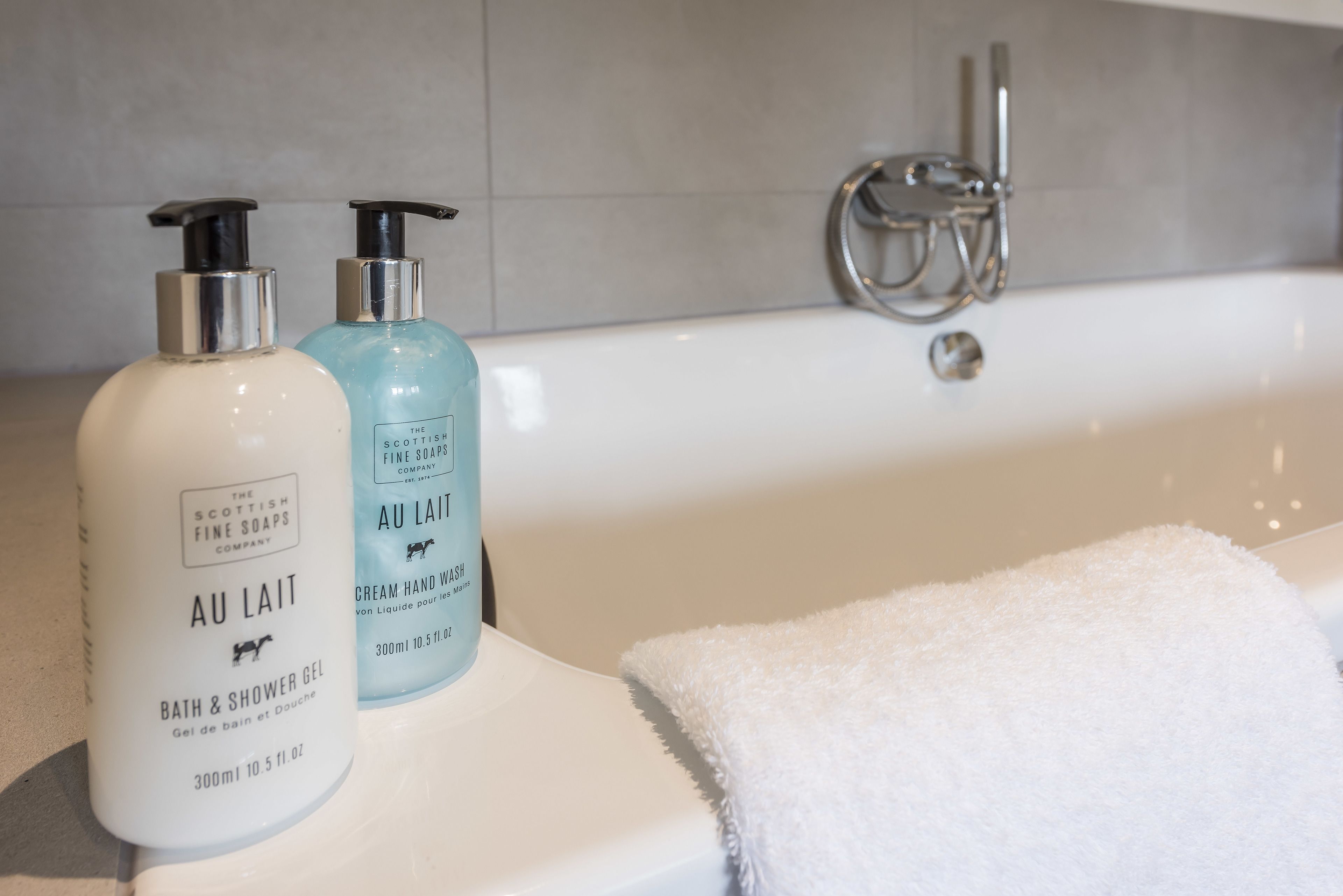 Two bottles of The Scottish Fine Soaps AU LAIT bath and shower gel and cream hand wash on a bathroom counter next to a white bathtub with a white towel.