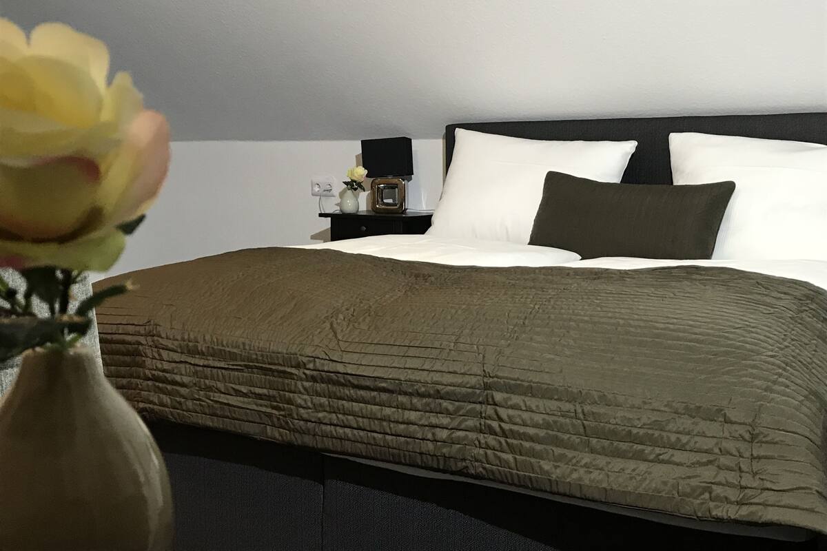 A neatly made bed with a dark headboard, white pillows, and a green blanket. There is a small bedside table with a vase of flowers and a lamp.