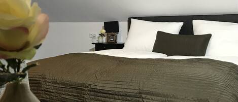 A neatly made bed with a dark headboard, white pillows, and a green blanket. There is a small bedside table with a vase of flowers and a lamp.