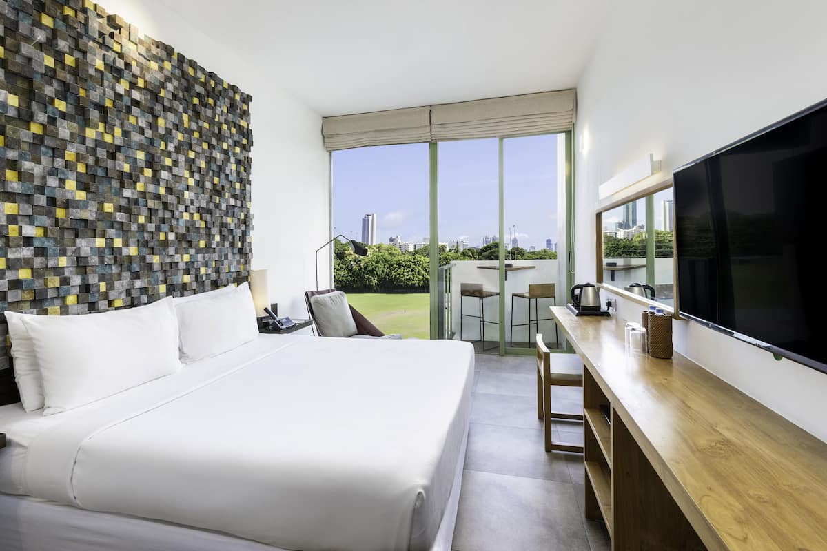 A modern hotel room with a large bed, a flat-screen TV, a wooden desk, and a balcony with a city view.