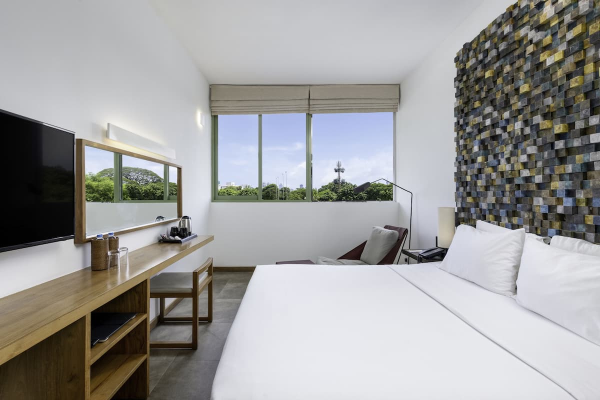 A modern hotel room with a large bed, a desk with a flat-screen TV, a window with a view of greenery, and a unique mosaic wall.